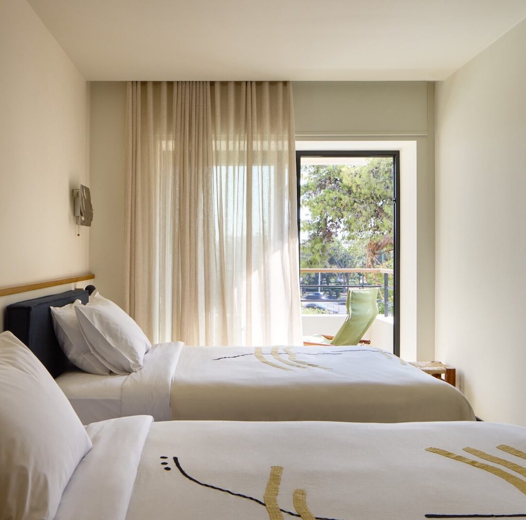 Two neatly made twin beds with white linens in a minimalist hotel room. Sunlight filters through curtains, highlighting a view of trees outside. A wall-mounted TV adds a modern touch.