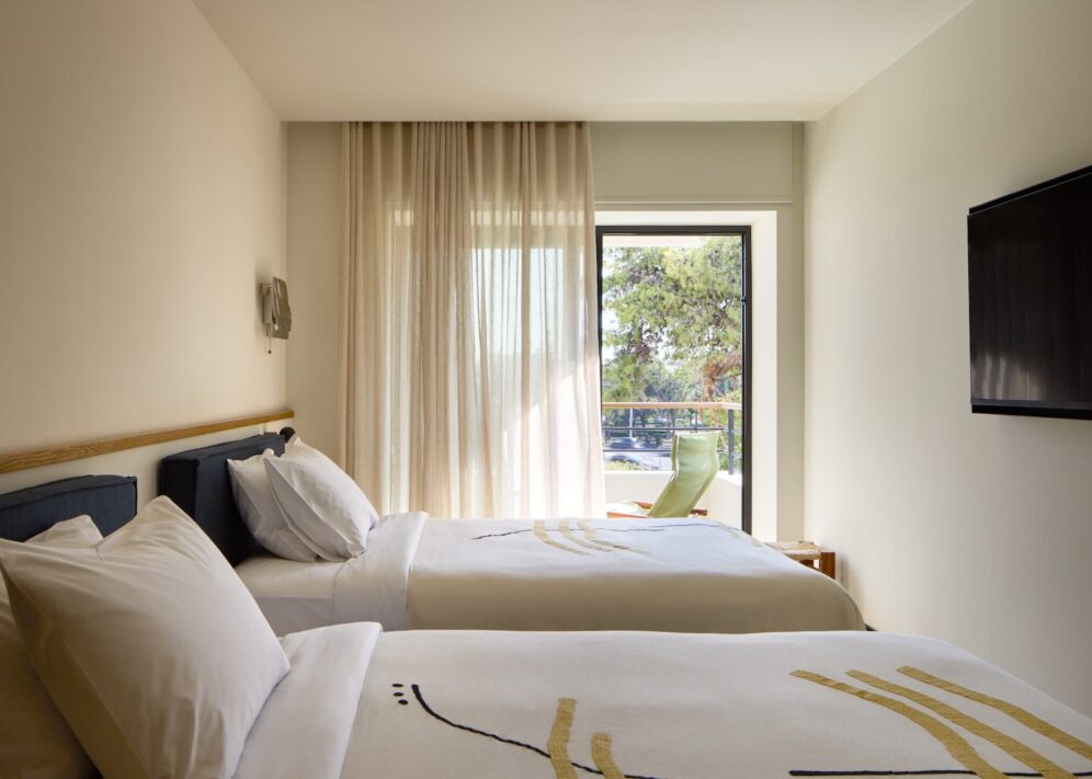 Two neatly made twin beds with white linens in a minimalist hotel room. Sunlight filters through curtains, highlighting a view of trees outside. A wall-mounted TV adds a modern touch.
