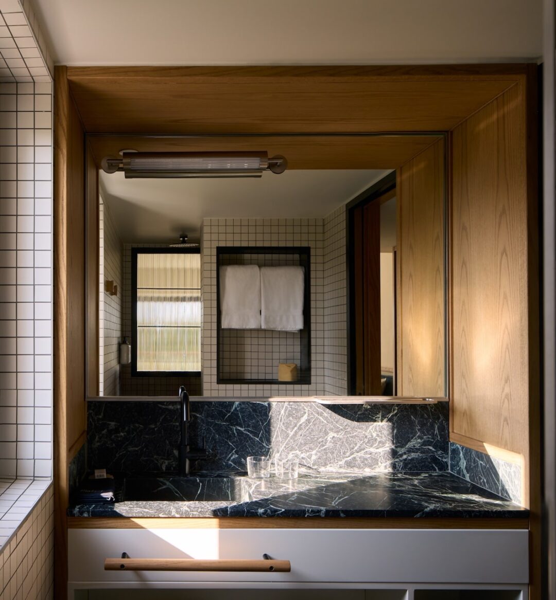 Modern bathroom with wooden accents and grid-patterned white tiles. Features a large mirror, black marble countertop, and neatly hung towels, conveying elegance and tranquility.
