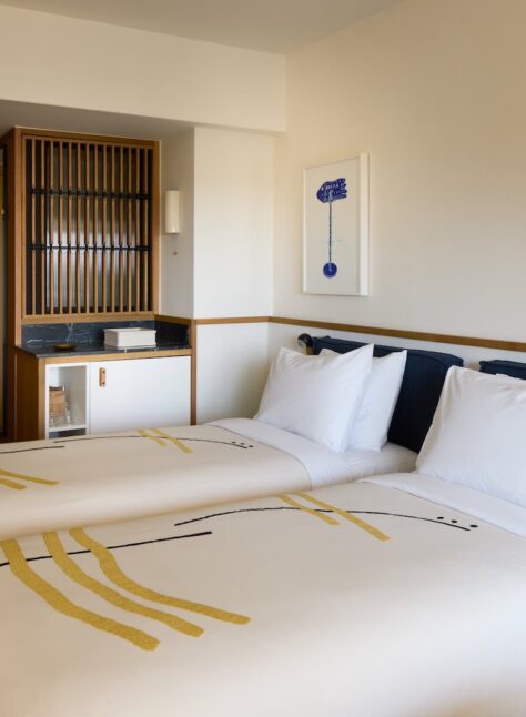 Modern hotel room with two neatly made single beds adorned with abstract-patterned blankets. Warm wooden accents, a small art piece, and soft lighting create a cozy atmosphere.