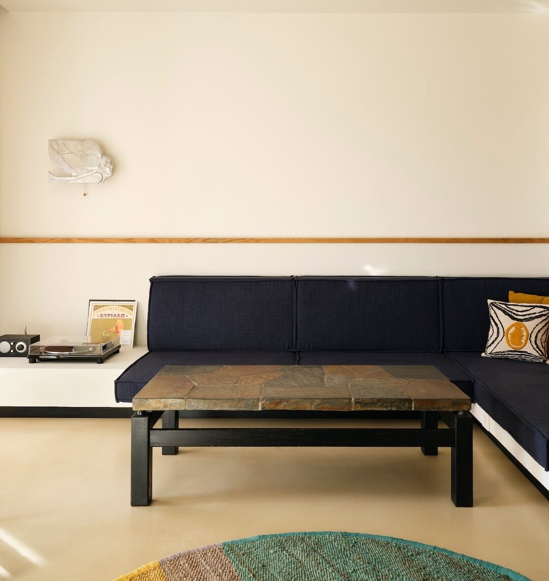 A minimalist living room with a navy blue L-shaped sofa, yellow and patterned cushions, a stone coffee table, and a vintage record player on a shelf.