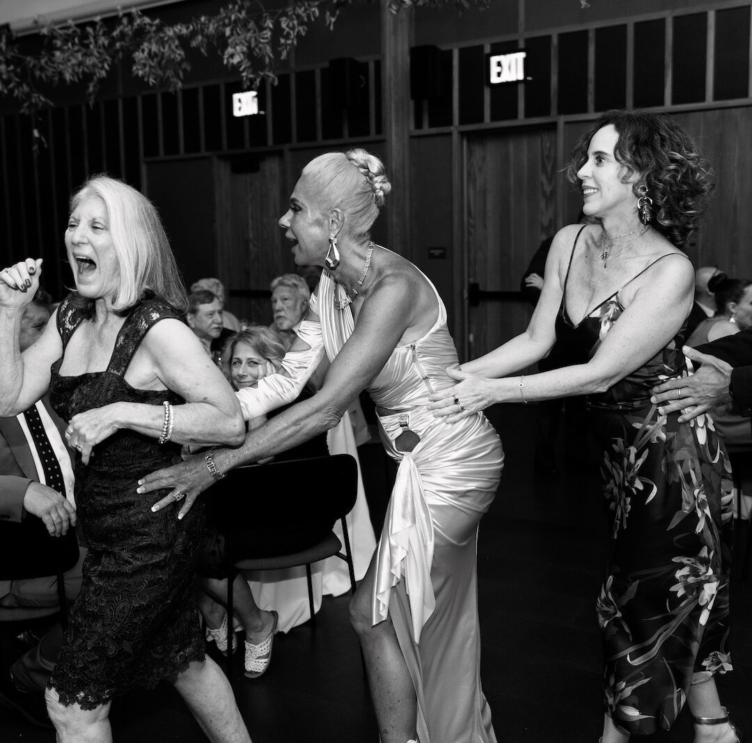 A group of people form a conga line and dance together at an indoor event, with tables and seated guests in the background.
