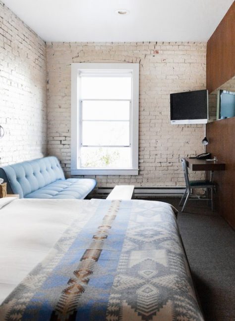 a room with exposed white brick walls is drenched in natural sunlight; a bed sits next to a long couch