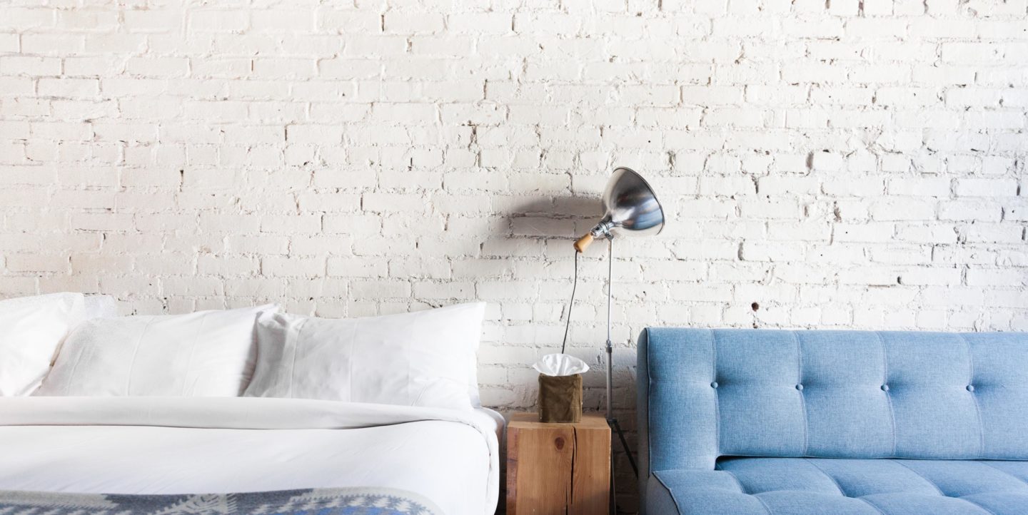 A hotel room with a bed and couch against a white brick wall