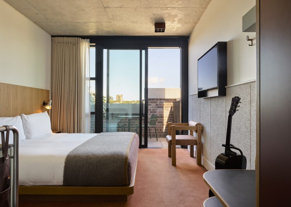 Modern hotel room with a double bed, TV, guitar, desk, and balcony with outdoor seating, featuring neutral tones and minimal decor.