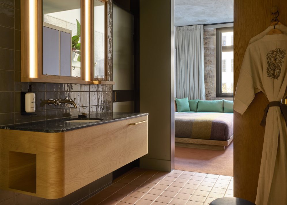 A sleek tiled wall supports to knobs that provide water to a black granite sink, and an entryway to the bed area is in the background