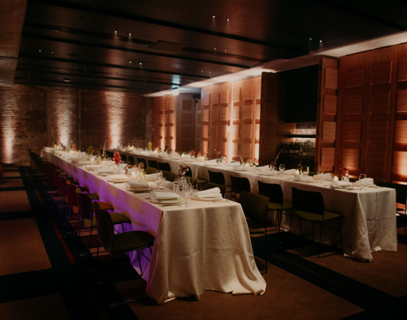 Two long banquet tables set for a formal dinner in a dimly lit, modern event space with brick and wood-paneled walls.