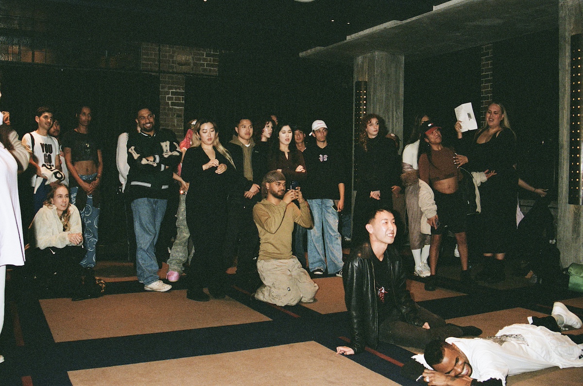 A diverse group of people, some seated and some standing, watch an event in a dimly lit room with brick walls and concrete pillars.
