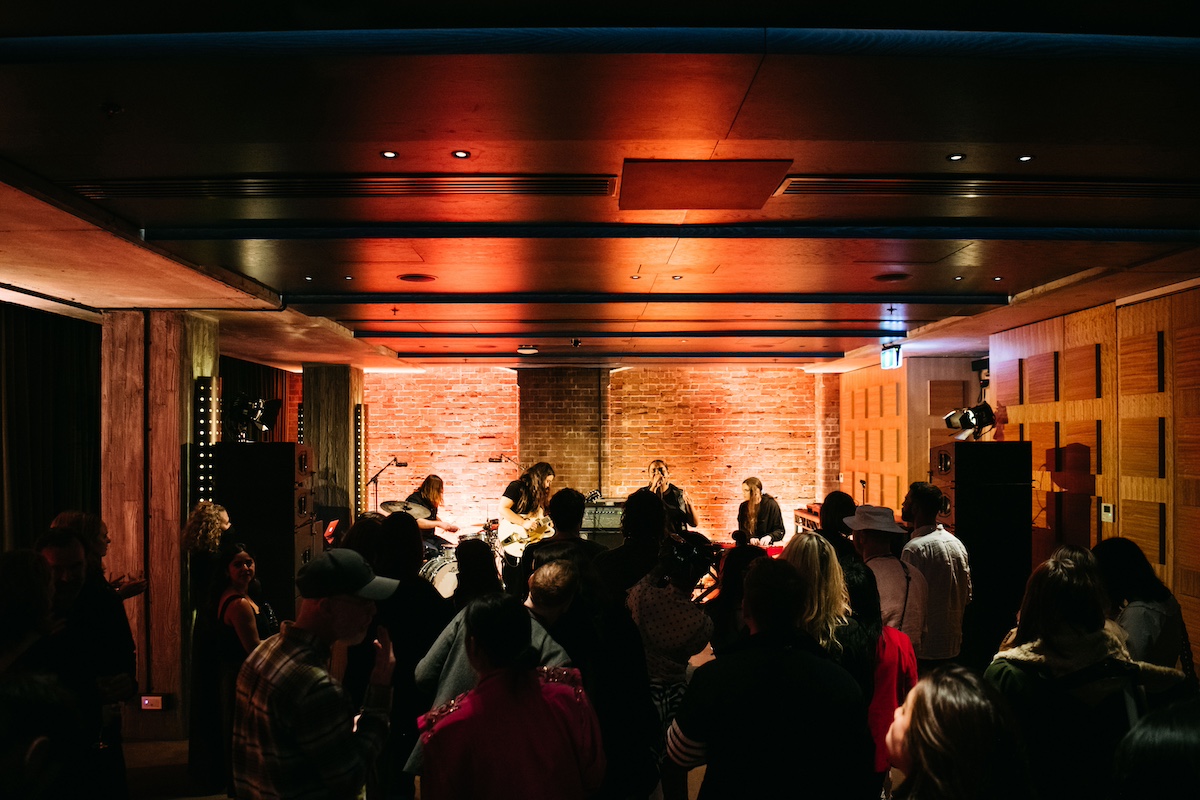 Crowd watches a live band perform on stage in a dimly lit venue with brick walls and wooden paneling.