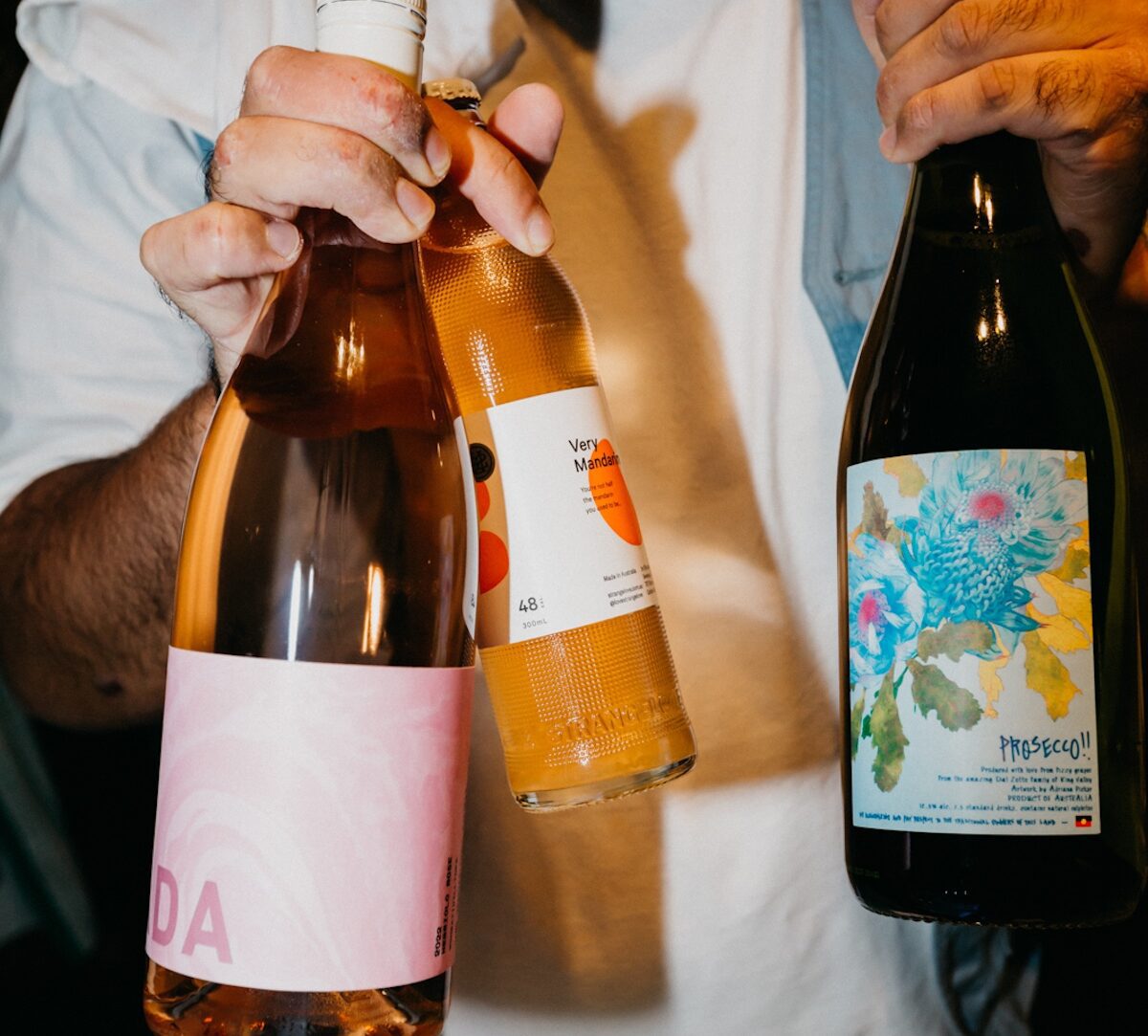 A person holds three bottles: one pink-labeled, one orange-labeled, and one with a floral design labeled “Prosecco!!”.